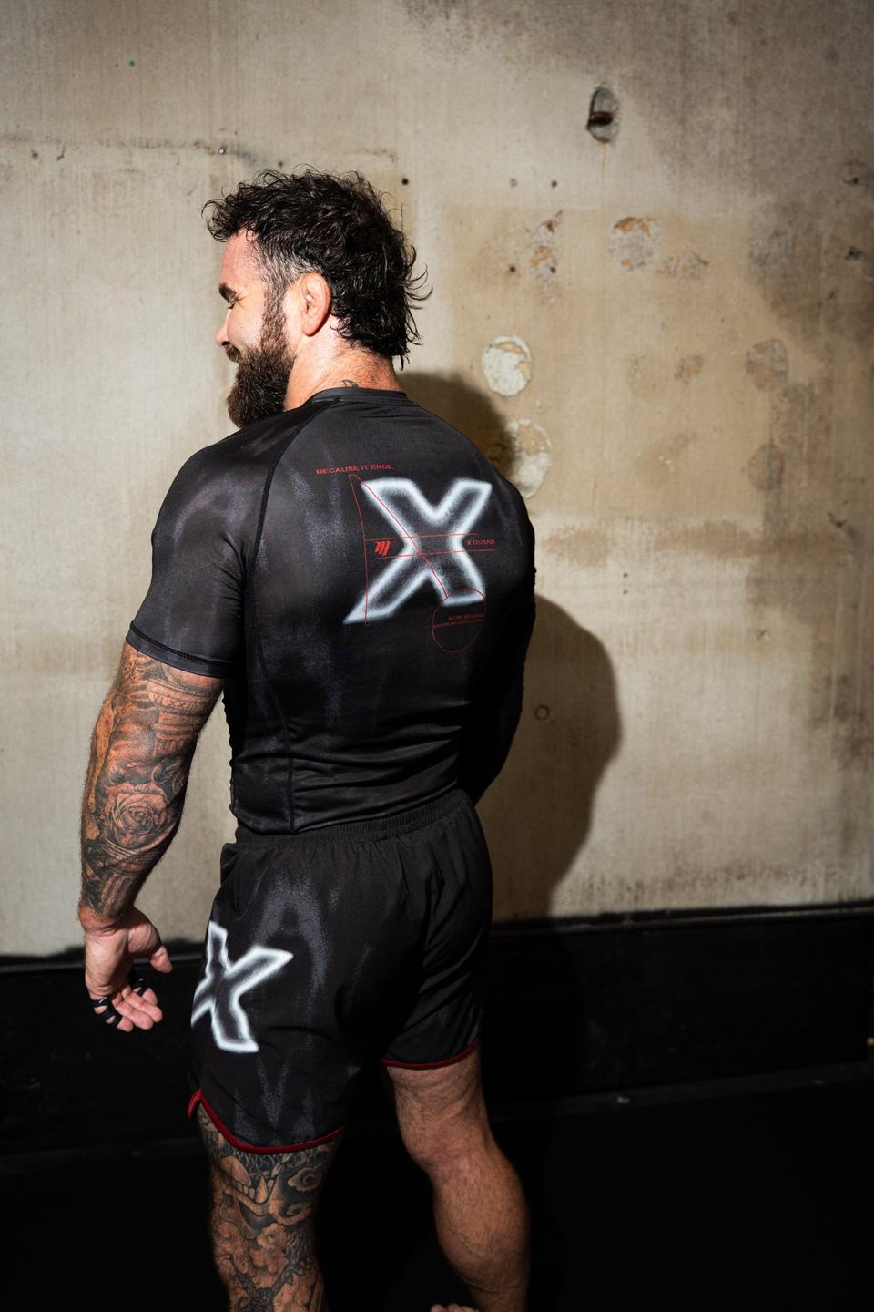 Cam Hurd from behind in a black X rashguard, facing the gym wall
