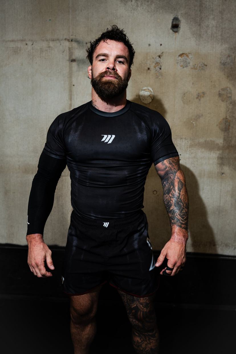 Cam Hurd in a black rashguard, standing square to the camera in front of a concrete wall